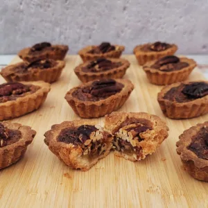 square photo with mini pecan tarts on a wooden board and one tart cut open to show inside