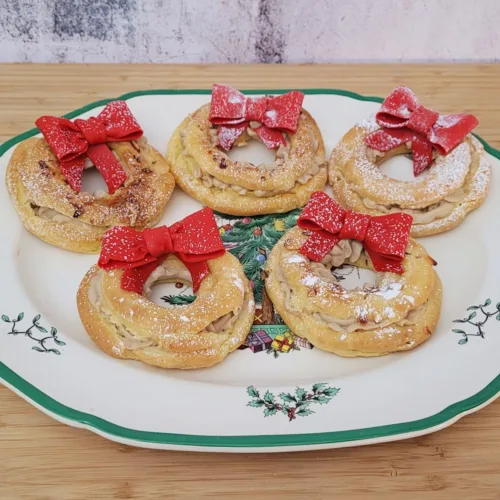 five cream puff wreaths with red bows on a plate and sprinkled with powdered sugar