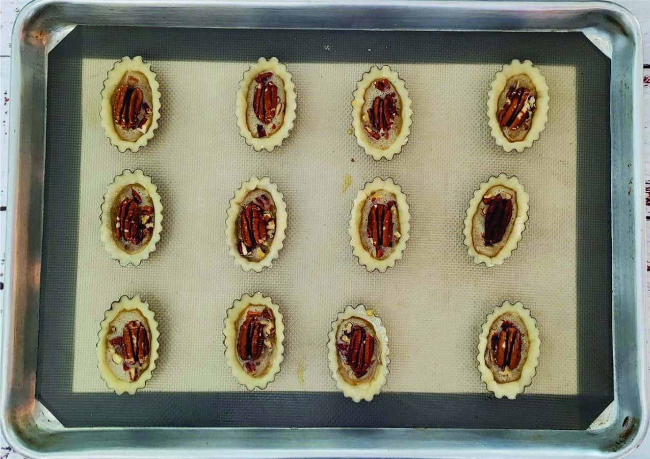 tart shells on a baking pan with pie filling and pecans in tarts