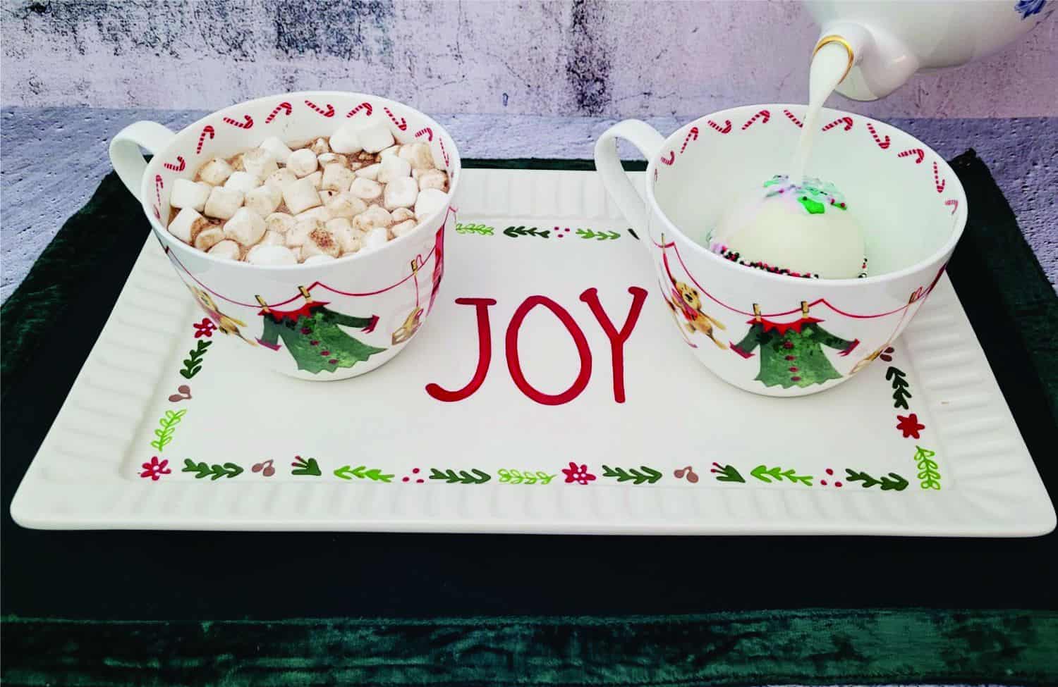 two Christmas mugs with cocoa bombs and hot milk being poured into mug