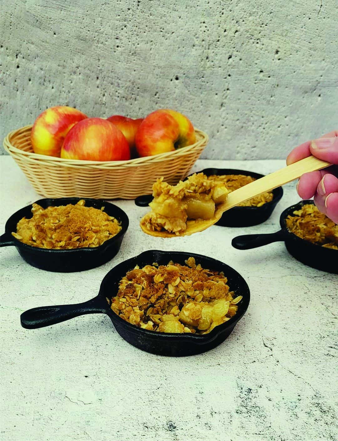 individual apples crisps in pans with a spoon digging into one pan. a basket of apples in the background