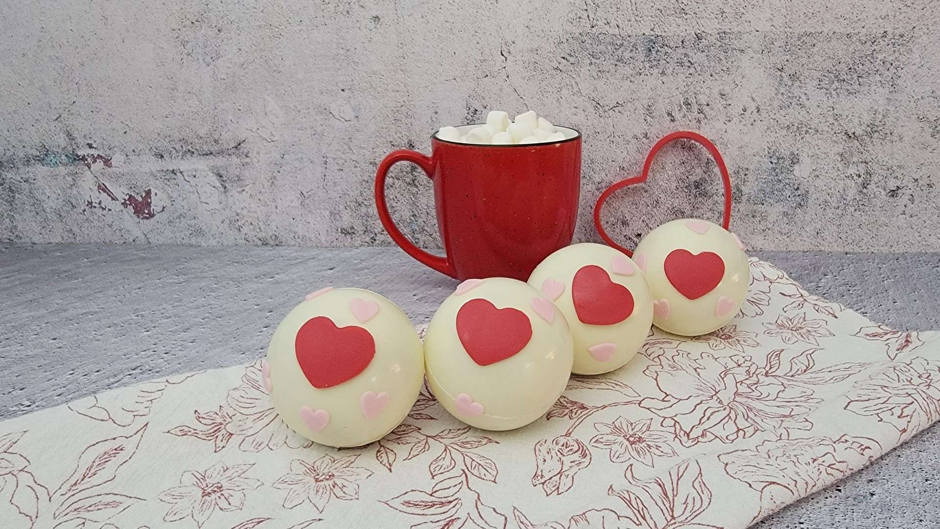 four hot chocolate bombs decorated with red and pink hearts and a red cup in the background