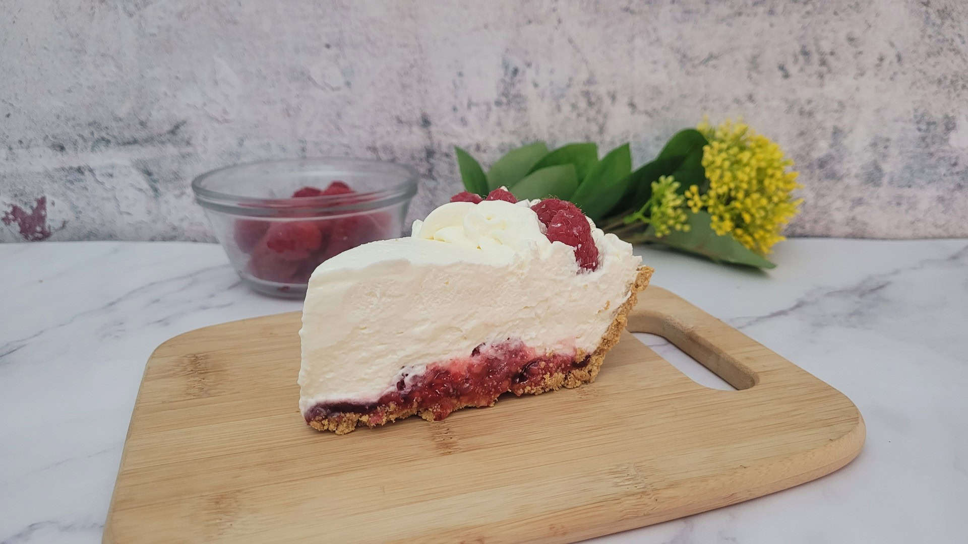 side view of a slice of no bake raspberry cream pie on a wooden board with a bowl of raspberries in the background