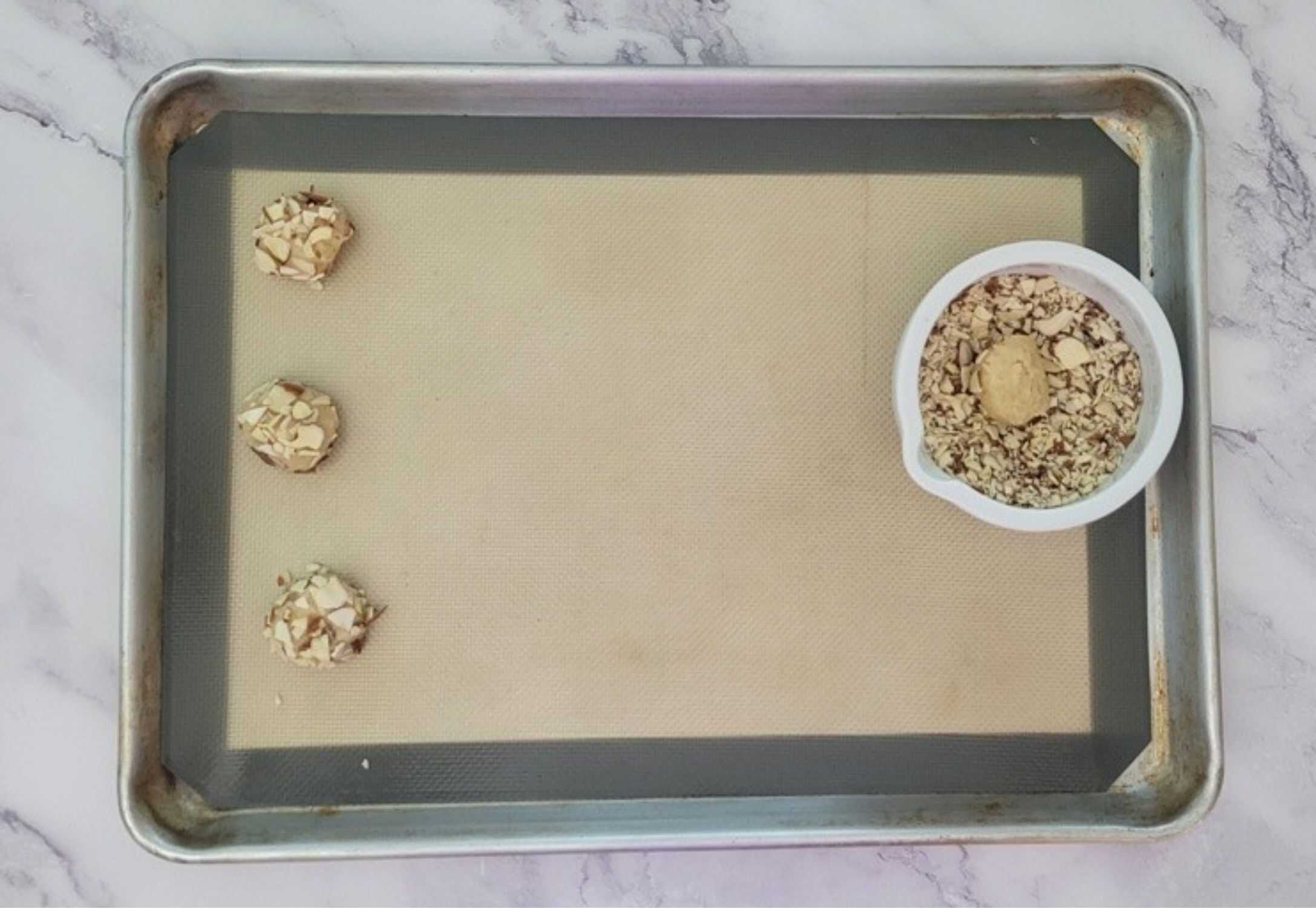 rolling chewy almond cookie dough into crushed almonds and placing on a baking sheet