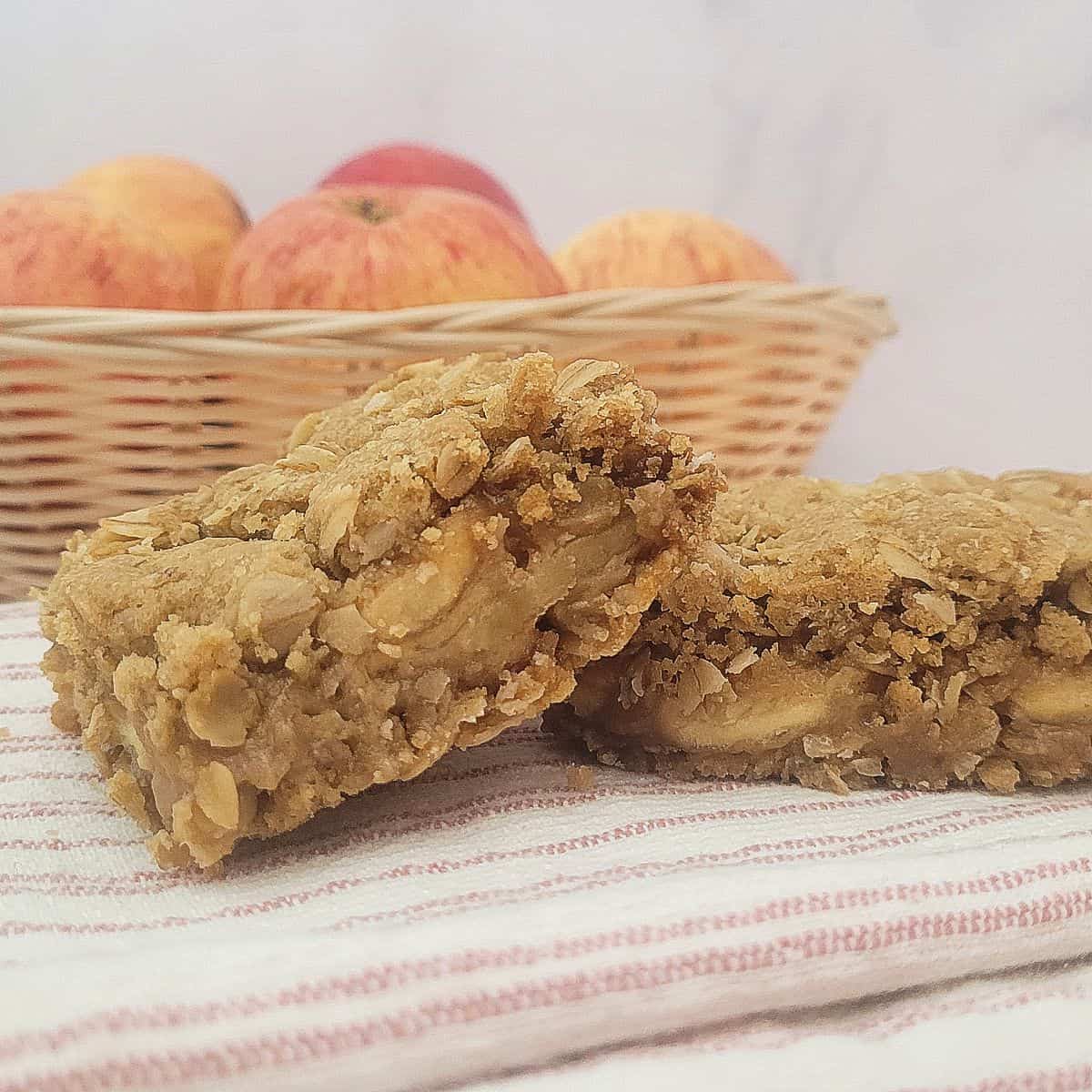 two slices of oatmeal apple bars on a napkin with a basket of apples in the background