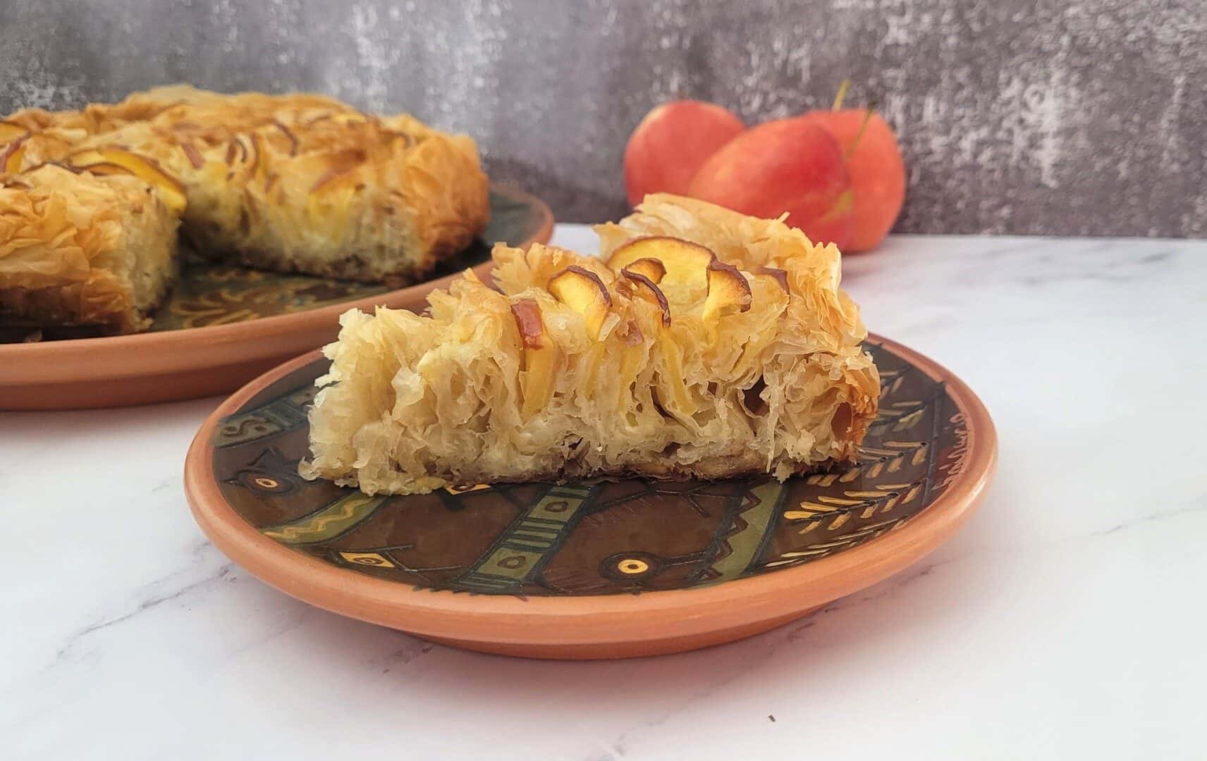 slice of apple cake made with phyllo dough and custard on a plate with remainder of cake in background