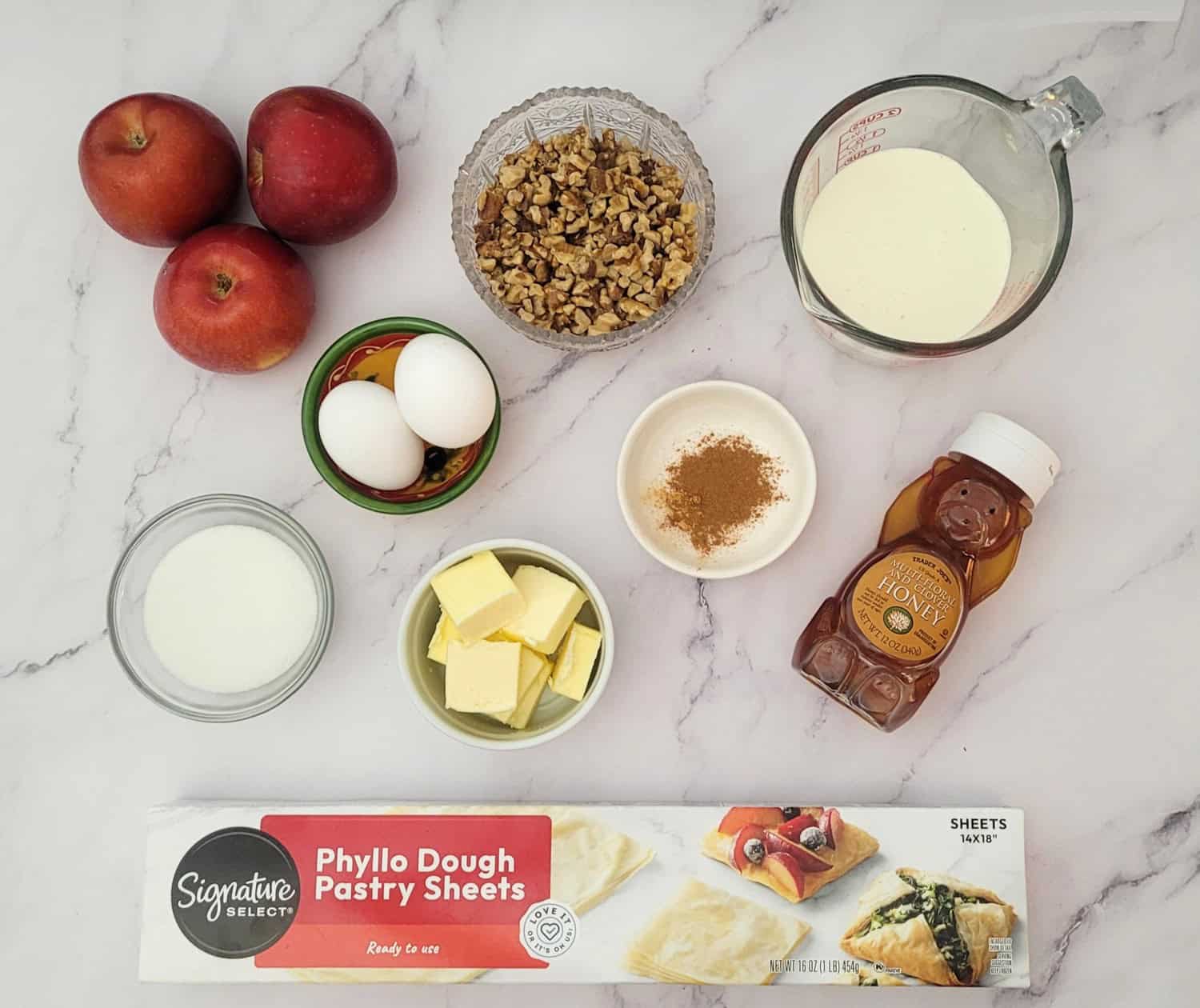 overhead view of ingredients to make crinkle apple cake including phyllo dough, honey, cinnamon, nutmeg, butter, granulated sugar, heavy cream, apples, walnuts and eggs