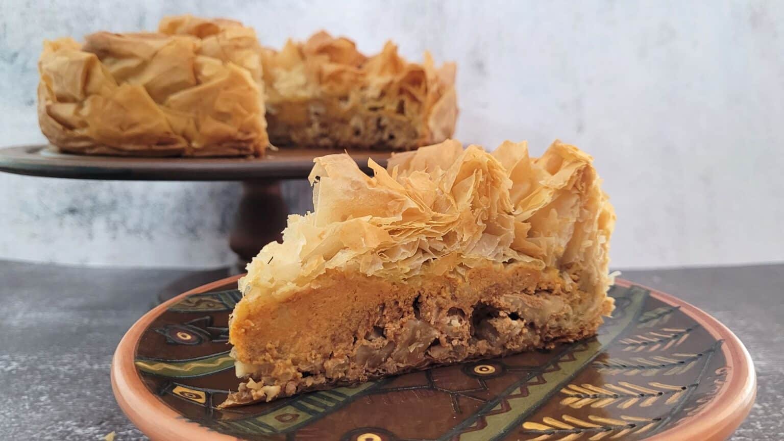 A close-up of a slice of layered pastry dessert on a decorative plate with intricate patterns, showcasing its flaky, golden-brown crust and a filling. The remaining dessert is on a stand in the blurred background. The scene is set against a simple light grey backdrop.