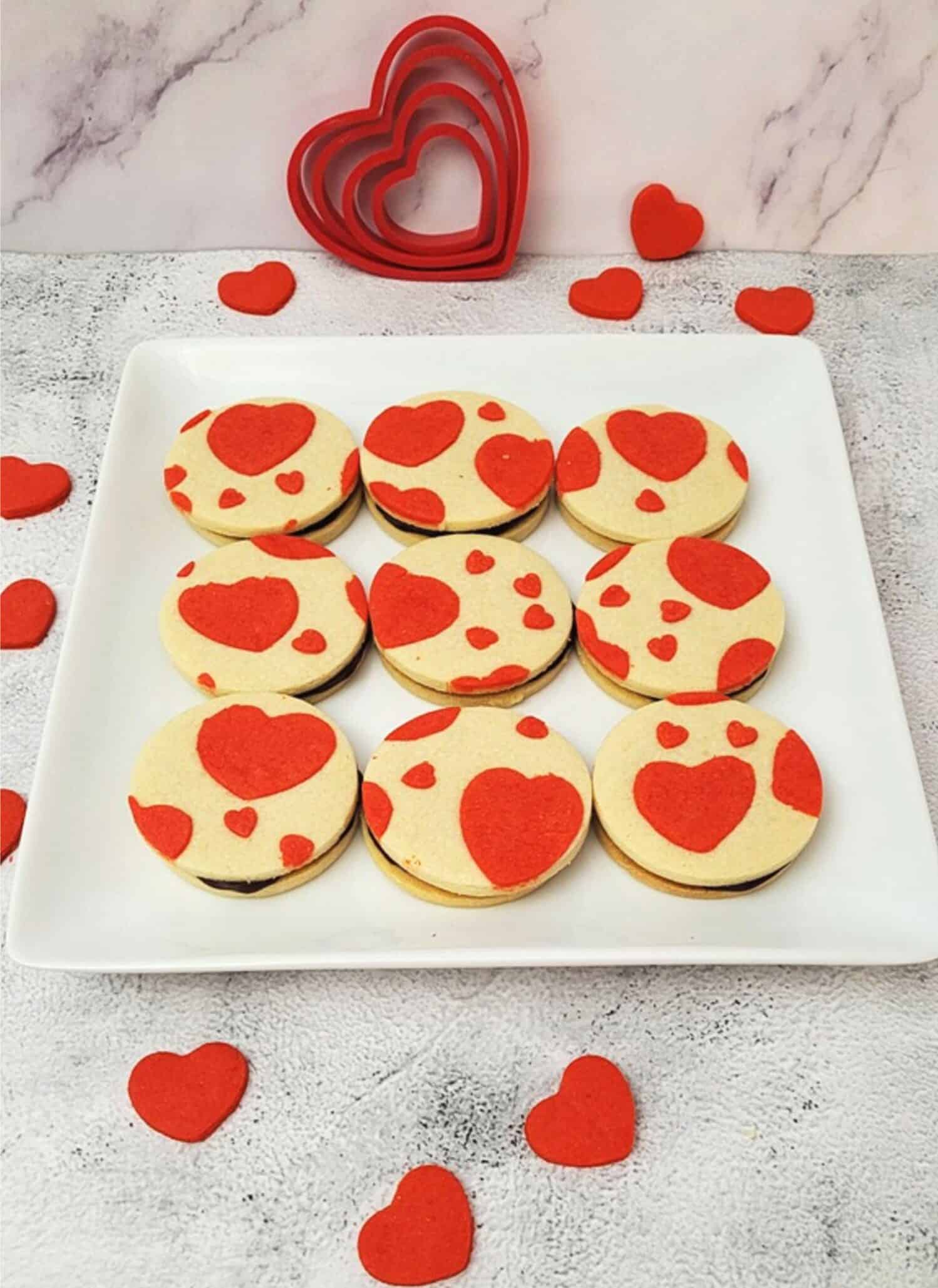 Valentine's heart sandwich cookies on a square serving platter with mini red heart cookies on countertop