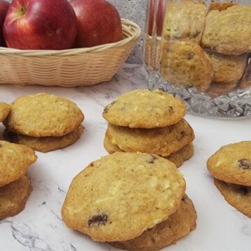 stacks of cookies on a countertop with cookie jar and basket of apples in the background