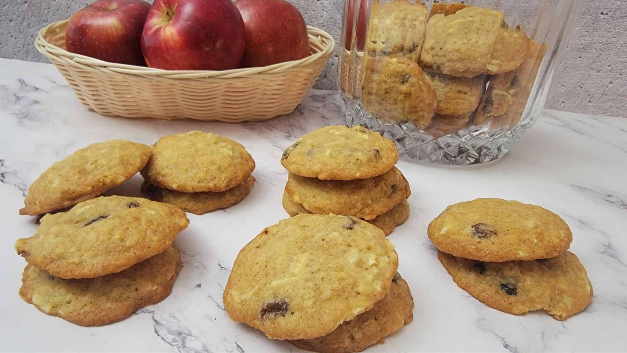 stacks of cookies on a countertop with cookie jar and basket of apples in the background